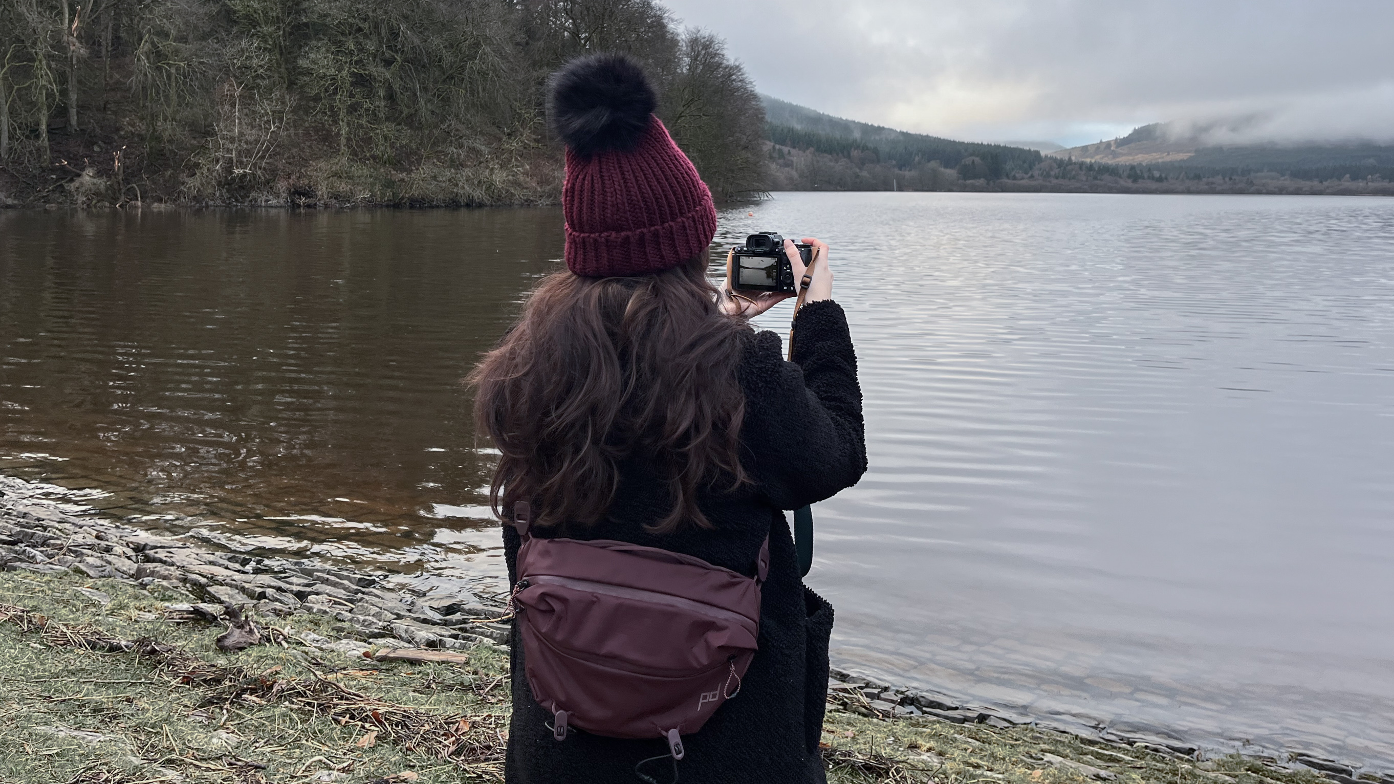 woman using a camera by a lake while wearing the Peak Design sling bag