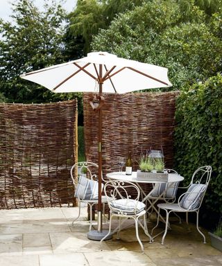 A natural wicker garden fencing idea behind a white parasol and white metal table and chairs.