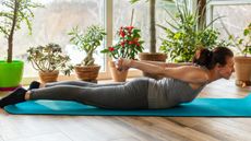 Woman lying on her front on a yoga mat with her chest lifted and hands clasped together above her butt. Pot plants line a floor-to-ceiling window behind her.