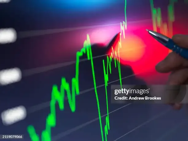 A hand holding a pen points to a bright stock market chart on a digital screen with fluctuating green and red graph lines indicating financial data 