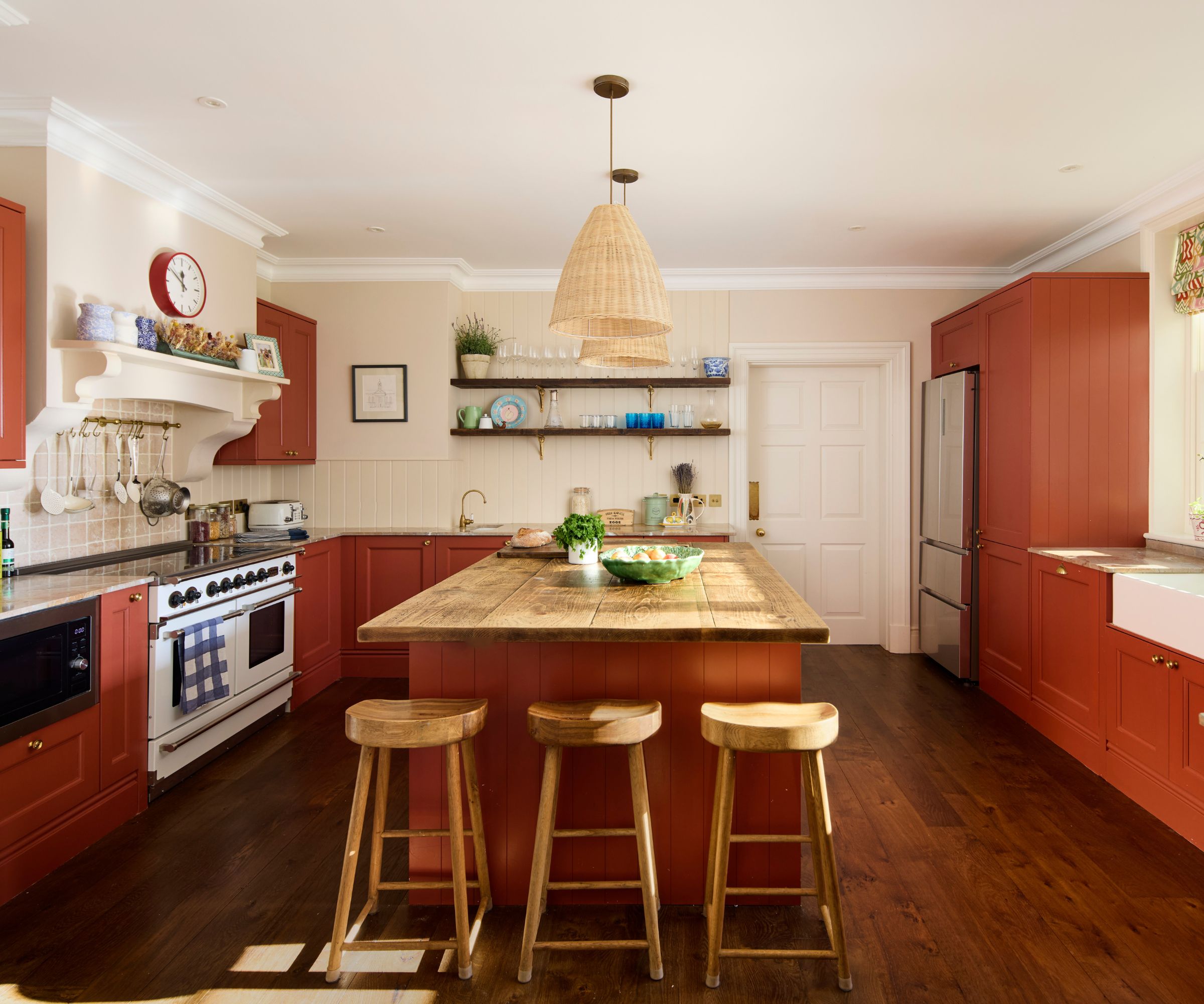 A soft kitchen with dark red cabinetry, warm white walls, wooden countertops, and open shelving displaying colorful glassware and decor