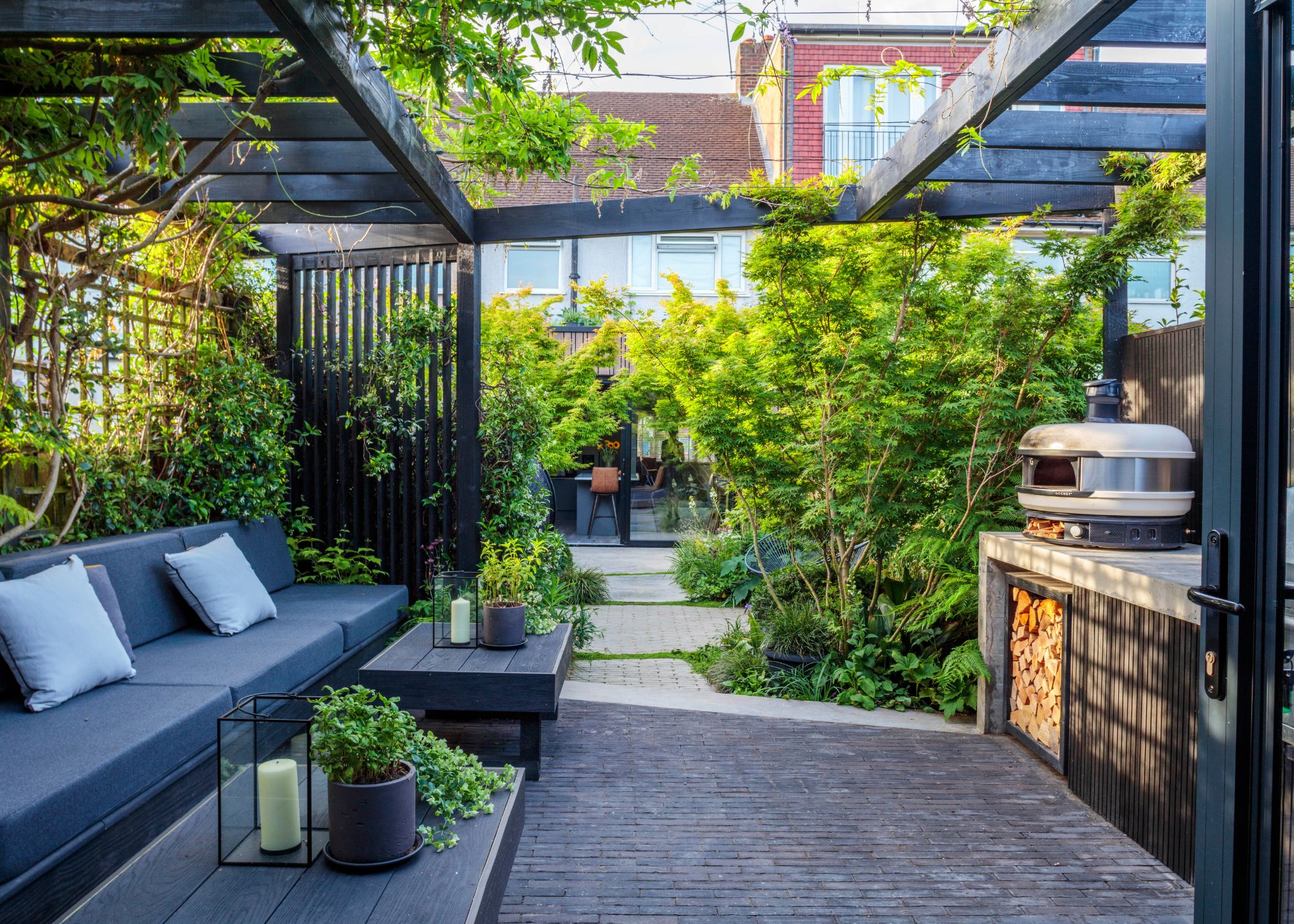An outdoor kitchen with seating, coffee tables and a counter with a pizza oven under a custom pergola surrounded by foliage