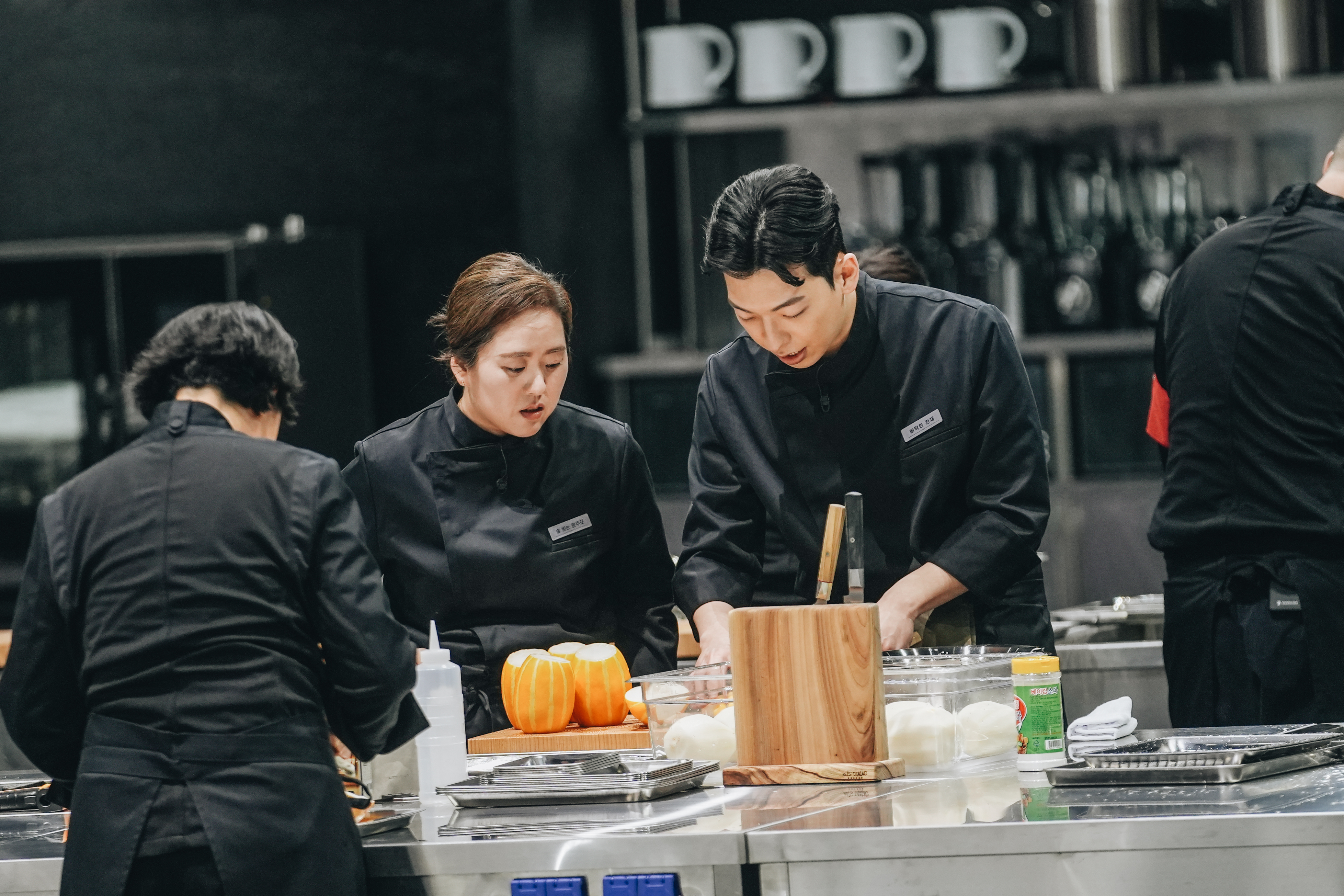 A female chef (Brewmaster Yun) and a male chef (Rebellious Genius) work over a cook station holding Korean melons and a wooden knife block. Other chefs dressed in black work around them, in 'Culinary Class Wars' season 2.