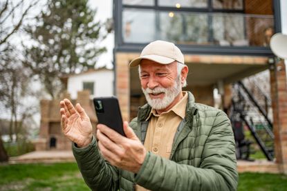 An elderly man stands in front of his cottage and uses a mobile phone to talk to his family