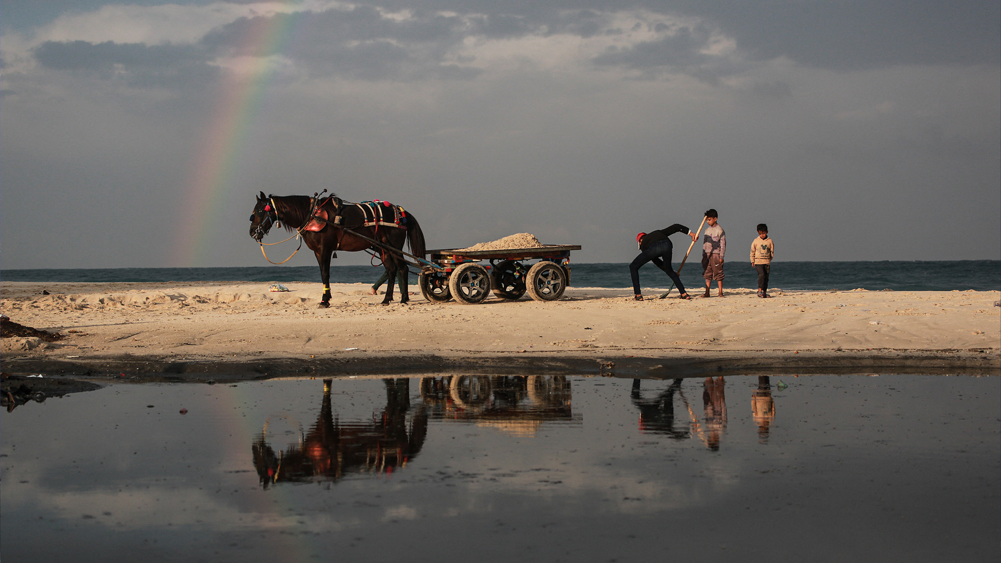 A man loads a horse cart with sand as a rainbow emerges over Deir al-Balah, in Gaza, Palestine