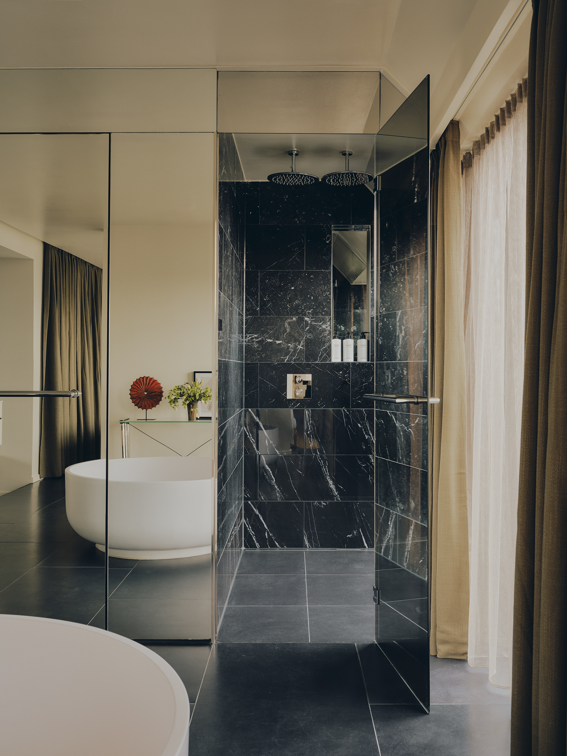 A hotel bedroom with a mirrored wardrobe that has a door open to reveal a black marble shower room inside next to a curtained window