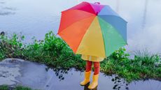 Little girl in yellow rubber boots with a rainbow umbrella standing in a large puddle
