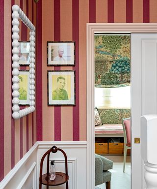 Light and dark pink striped wallpaper, white bobbin style mirror, artwork, white paneling, wooden side table, looking through to room with green and blue patterned blind