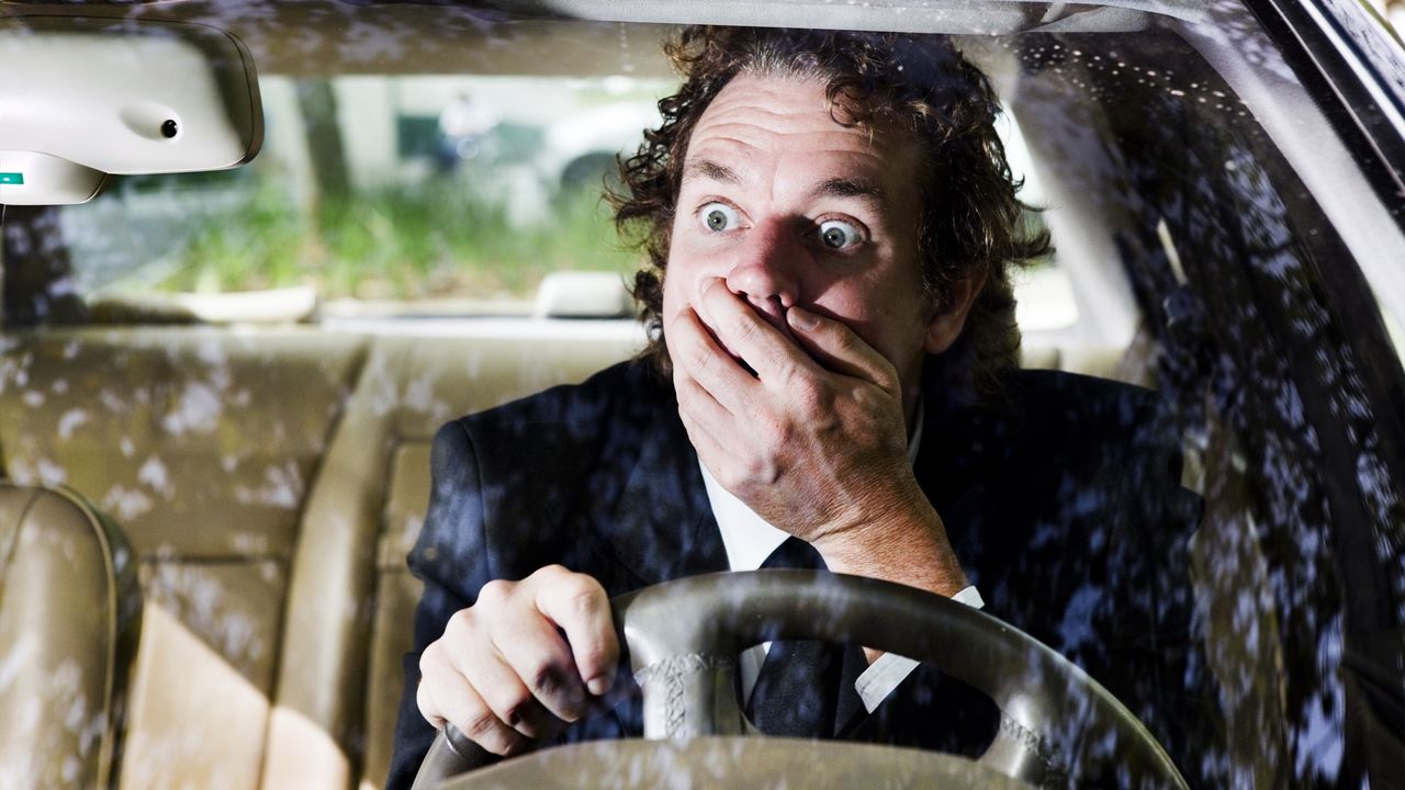 A man behind the wheel of a car covers his mouth and looks shocked.