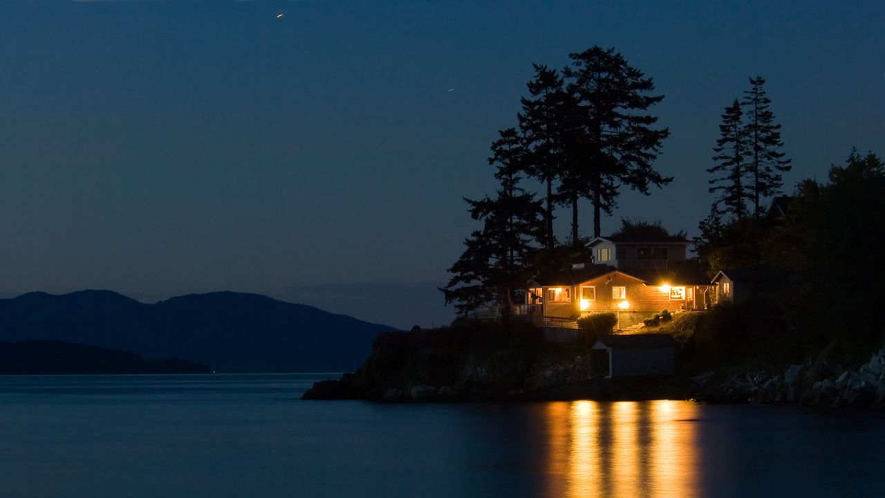 A lakeside home is lit up on night, its lights reflecting on the surface of the water.