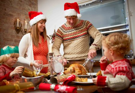 Family sitting at dinner table with father carving Christmas turkey. Parents wearing Christmas hats.