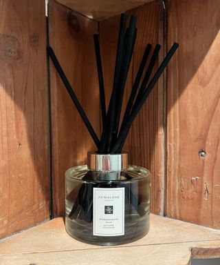 A Jo Malone pomegranate noir reed diffuser with black reeds on an antique wooden shelf