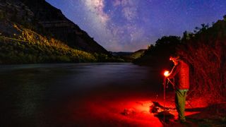 Photographer at Night Capturing Astrophotography Milky Way Landscape - Man with headlamp photographing a bright galaxy along scenic canyon and river with dark skies.