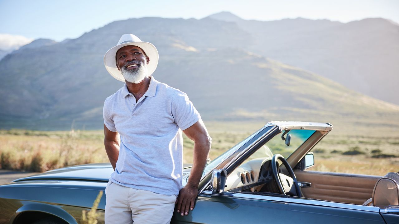 An older man leans against his convertible and smiles as he takes in mountainous scenery.