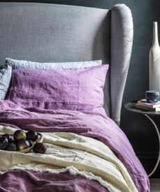 A bowl of figs on purple sheets on a gray headboard against a gray wall.