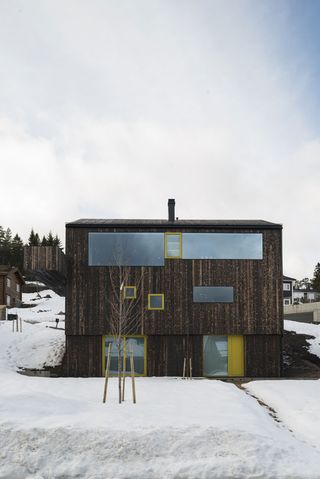 Alternative exterior view of the Oslo family house by STA during the day - the house features a dark wood exterior and windows and doors with yellow frames. There is snow on the ground and other houses nearby
