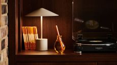 A yellow glass reed diffuser on a wood shelf near a portable white lamp, some books, and a record player