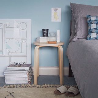 A wooden side table in a bedroom with blue walls, with a bed on one side and a pile of books on the other.