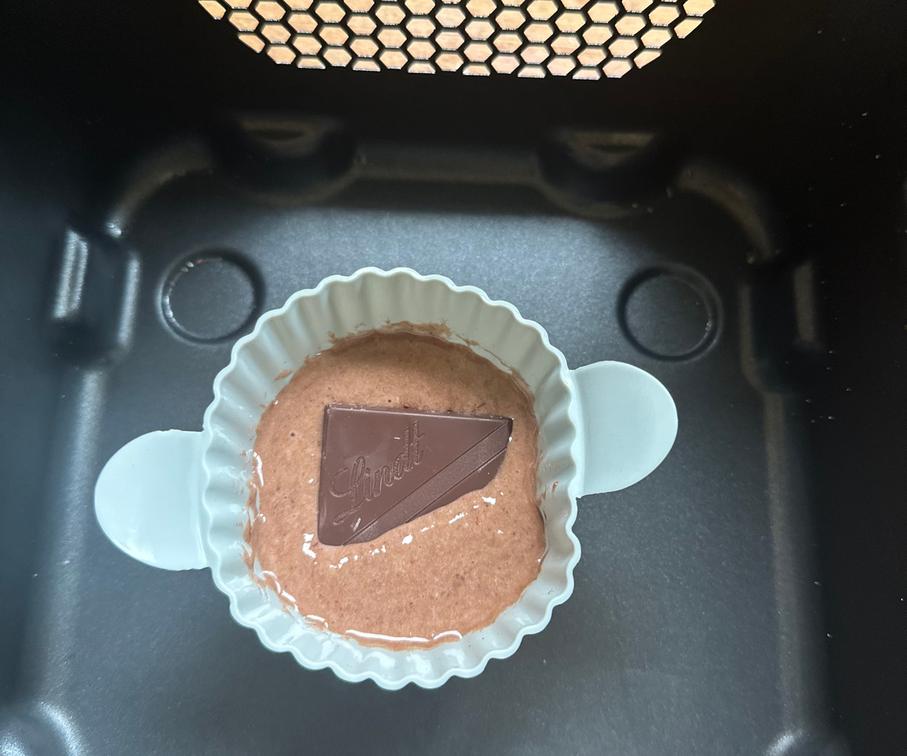 A small round blue silicone baking ramekin with chocolate cake batter and a square of chocolate inside, in a black air fryer basket