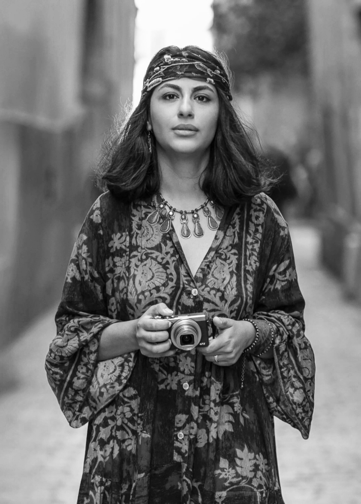 A woman in a floral patterned robe holds a camera, standing in a narrow street bathed in soft light