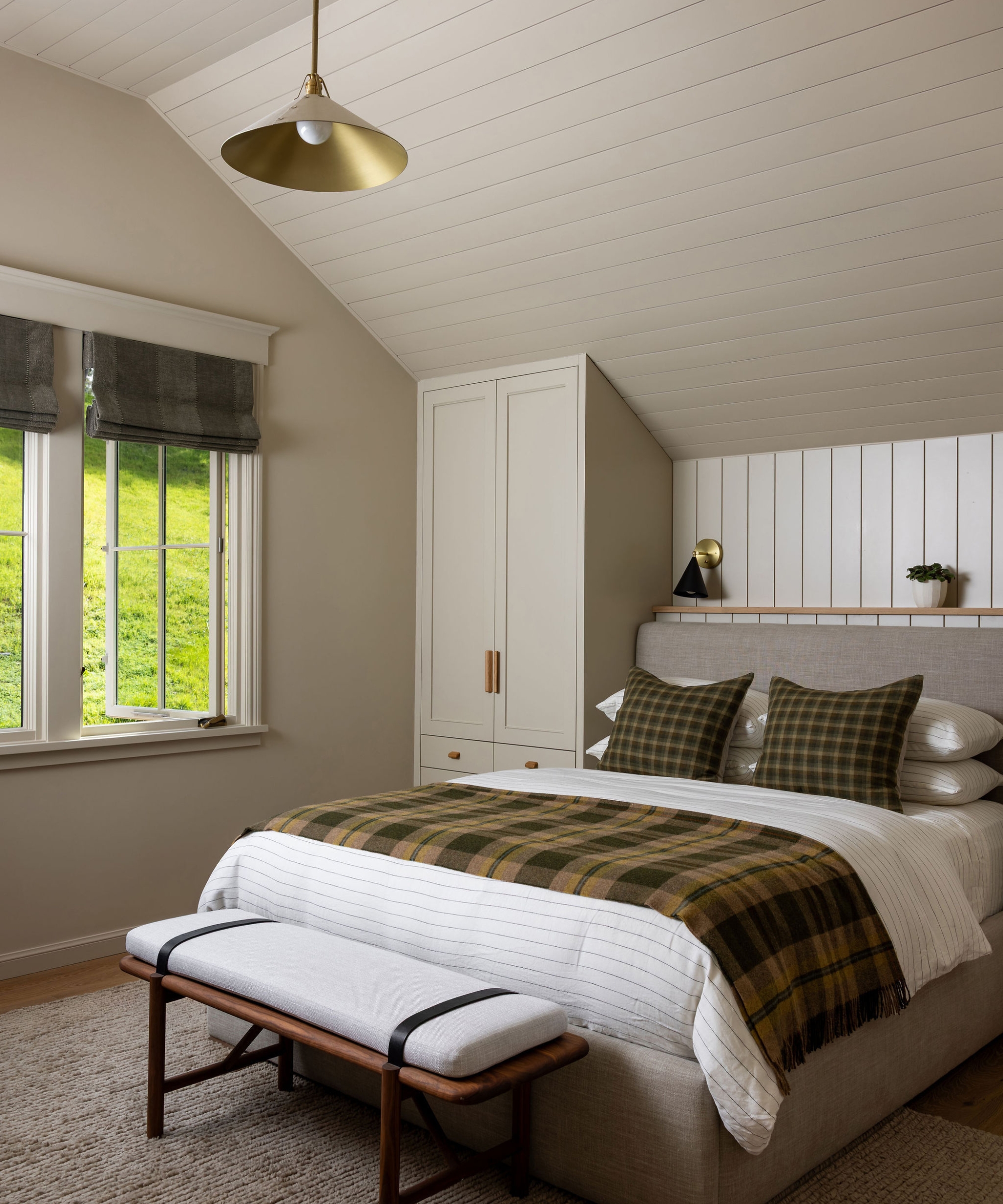 Boy’s bedroom with a sloped paneled ceiling, upholstered bed, green plaid textiles, built-in wardrobe and cushioned wood bench.