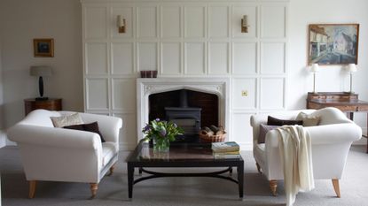 Bright white living room area featuring a pair of white antique-style sofas, a glass-top coffee table, and shaded wall sconces flanking the fireplace