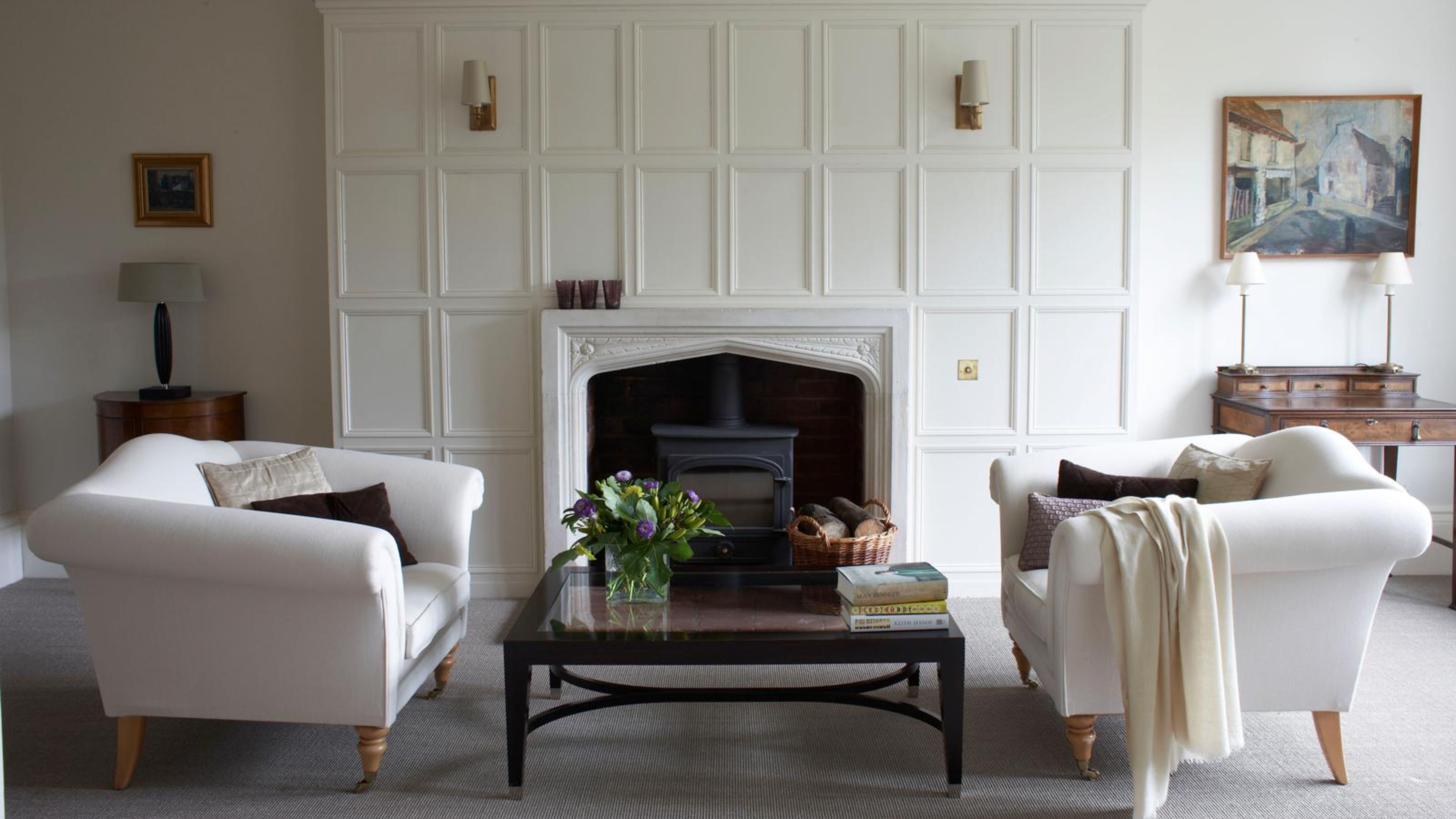 Bright white living room area featuring a pair of white antique-style sofas, a glass-top coffee table, and shaded wall sconces flanking the fireplace