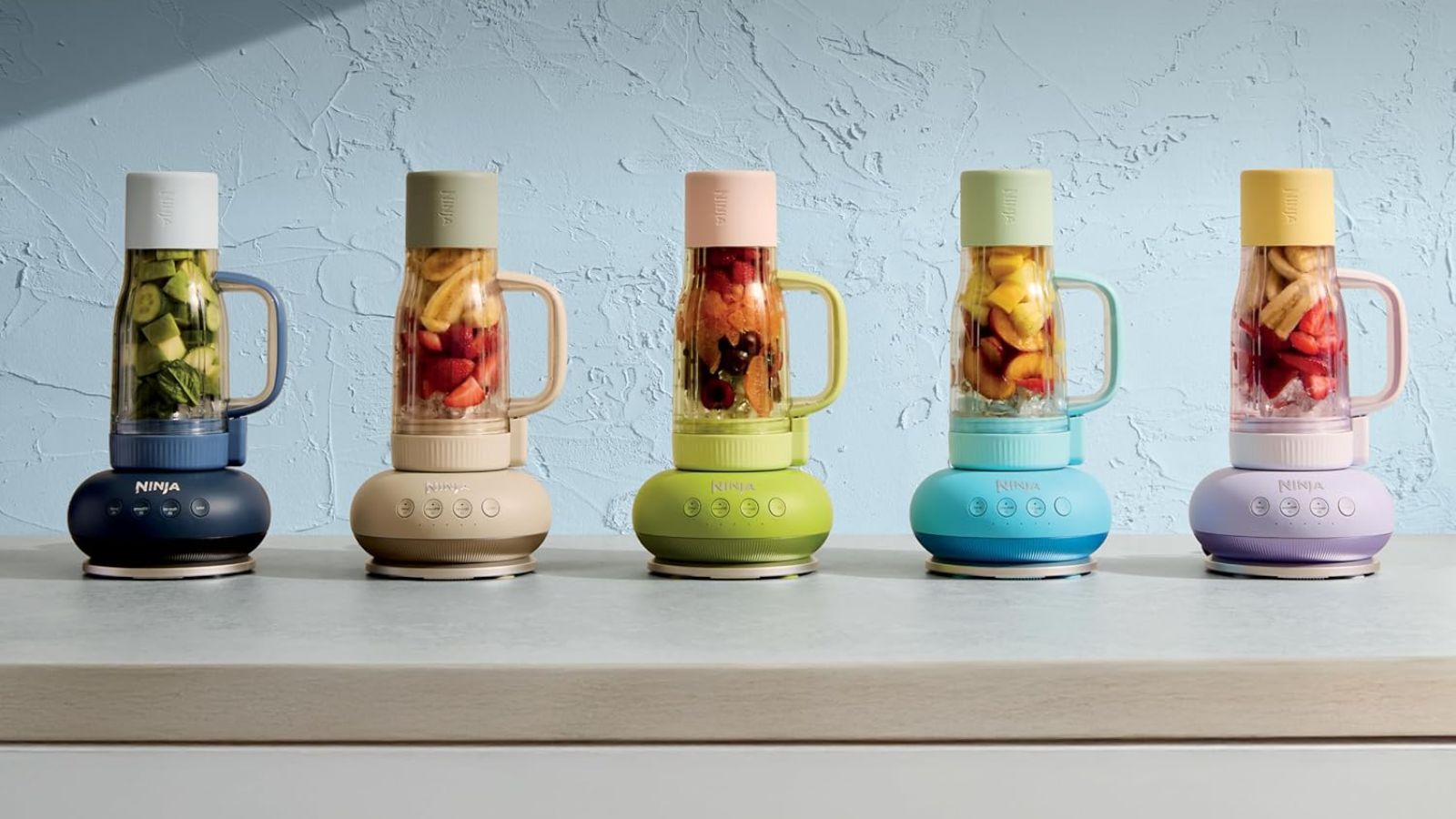 A colourful set of five tumble style drinking cups, upside down on blender bases, against a blue wall