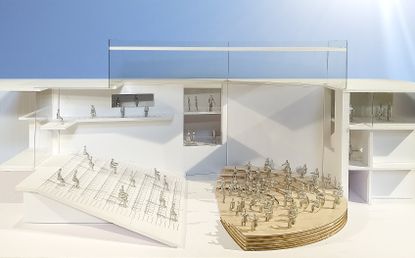 A model of the white Interior view of the center showing the auditorium with gold semi circle platform on the right, and the theatre-like seating area on the left. Silver human shaped objects placed on the seating area and the stage. 