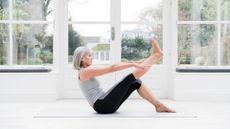 Woman in her 60s with grey bobbed hair stretching leg at home in front of windows overlooking a garden.