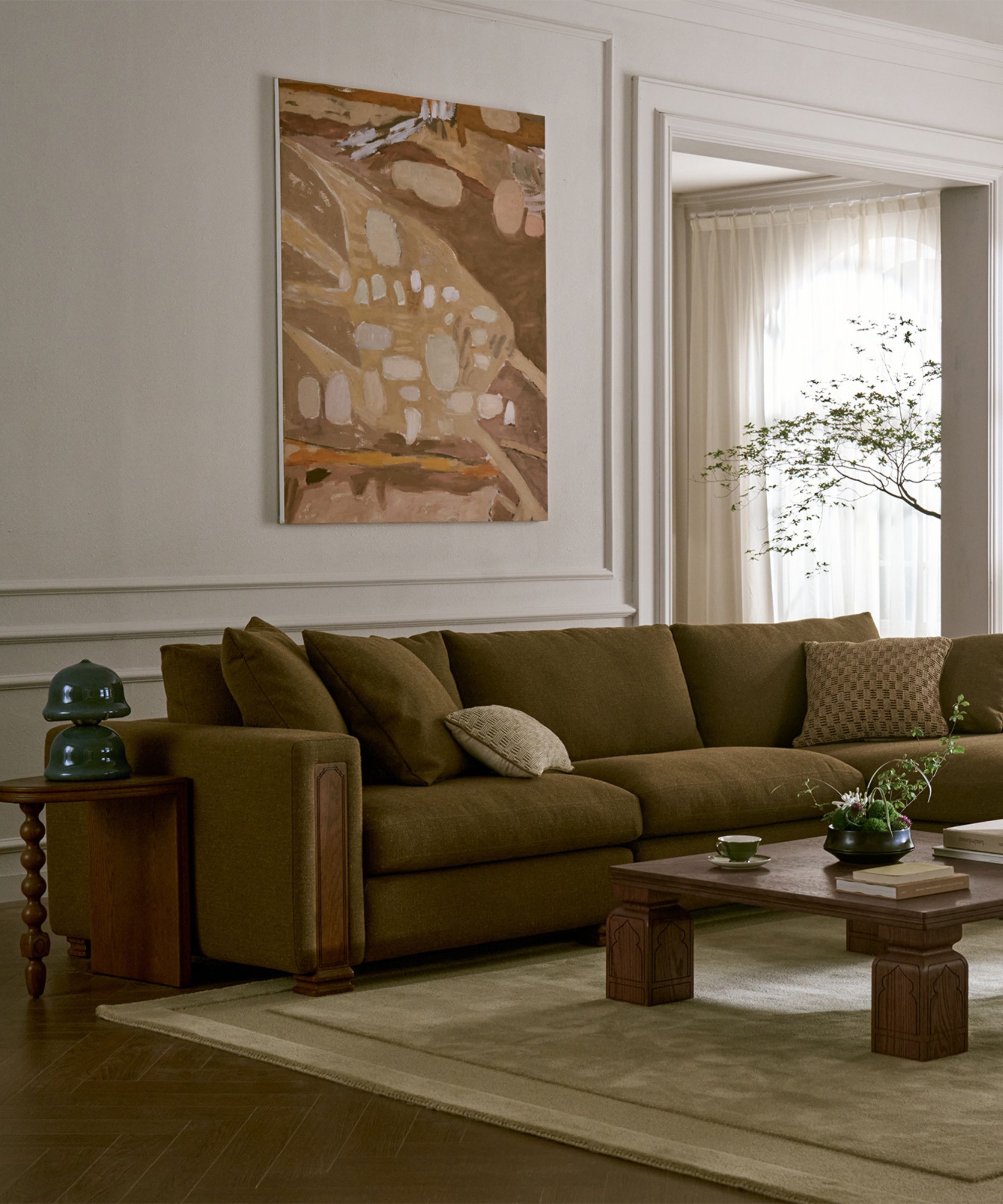 a living room with Castlery x Tan France furniture including an olive sofa with timber detailing, a timber coffee table styled with books and vases, a large artwork hung above, and a side table