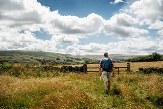 A man with a bagpack walking through tall grass, with a beautiful landscape in the background.