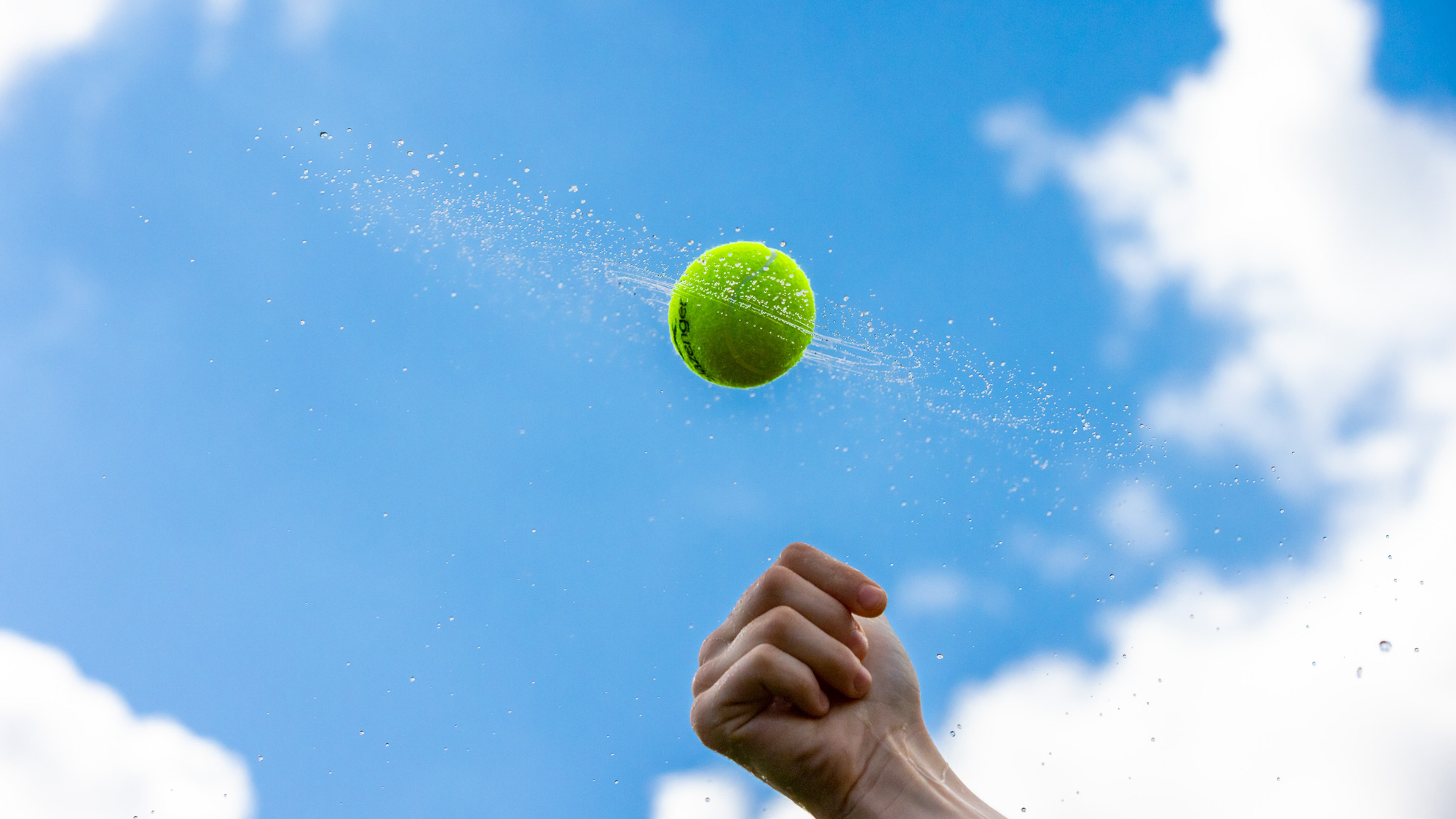 A wet tennis ball is spun against a bright blue sky which has lost definition in the water droplets