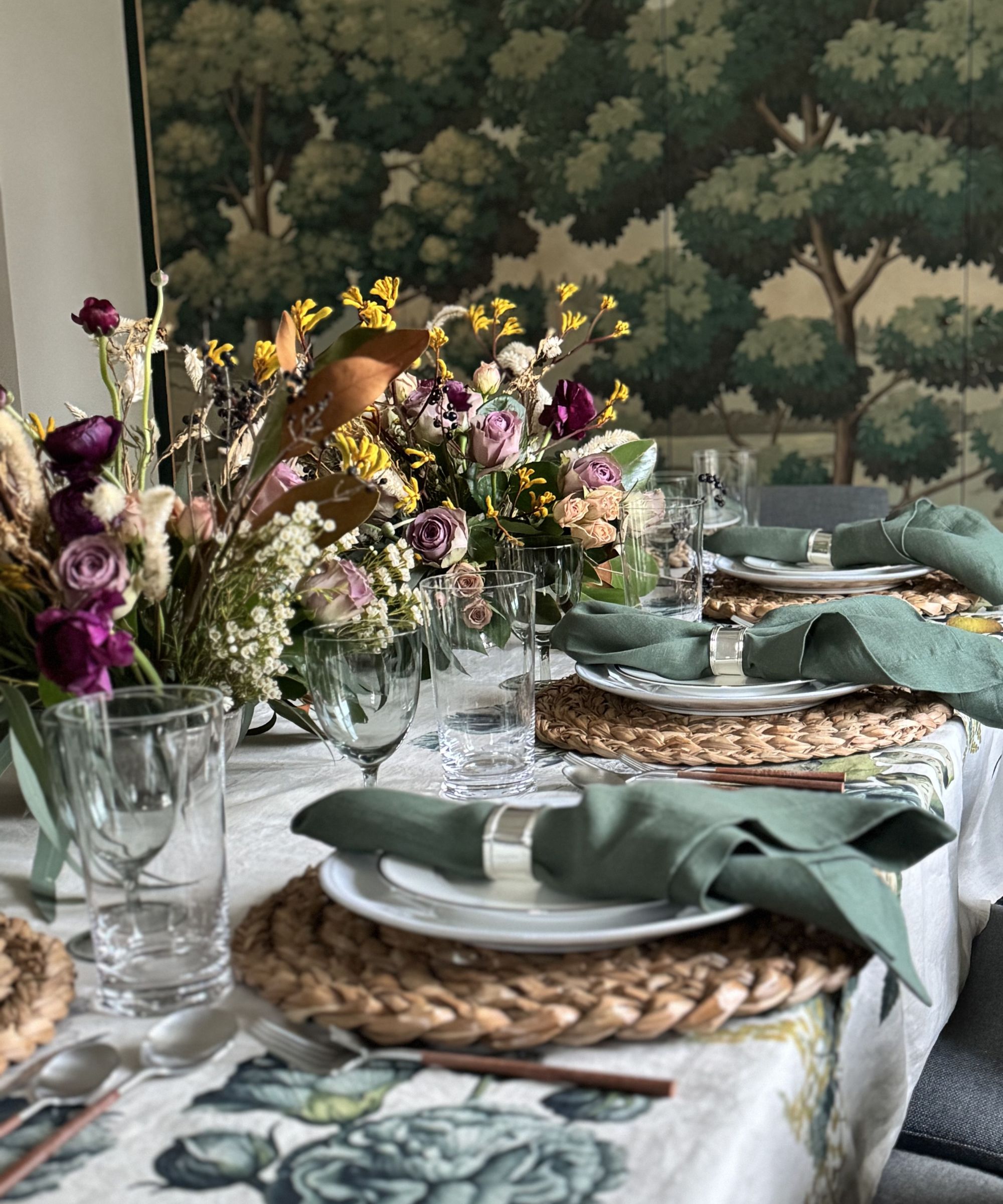 green themed table laid for thanksgiving with a white and green tablecloth, fall foliage centerpiece, wicker platemats and little velvet pumpkins
