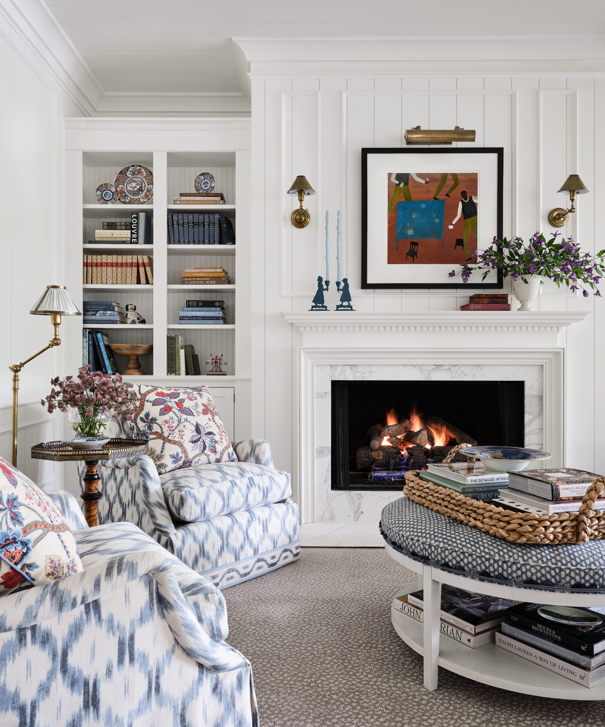 Family room with white paneled walls, a marble fireplace, blue ikat armchairs and a round upholstered ottoman.