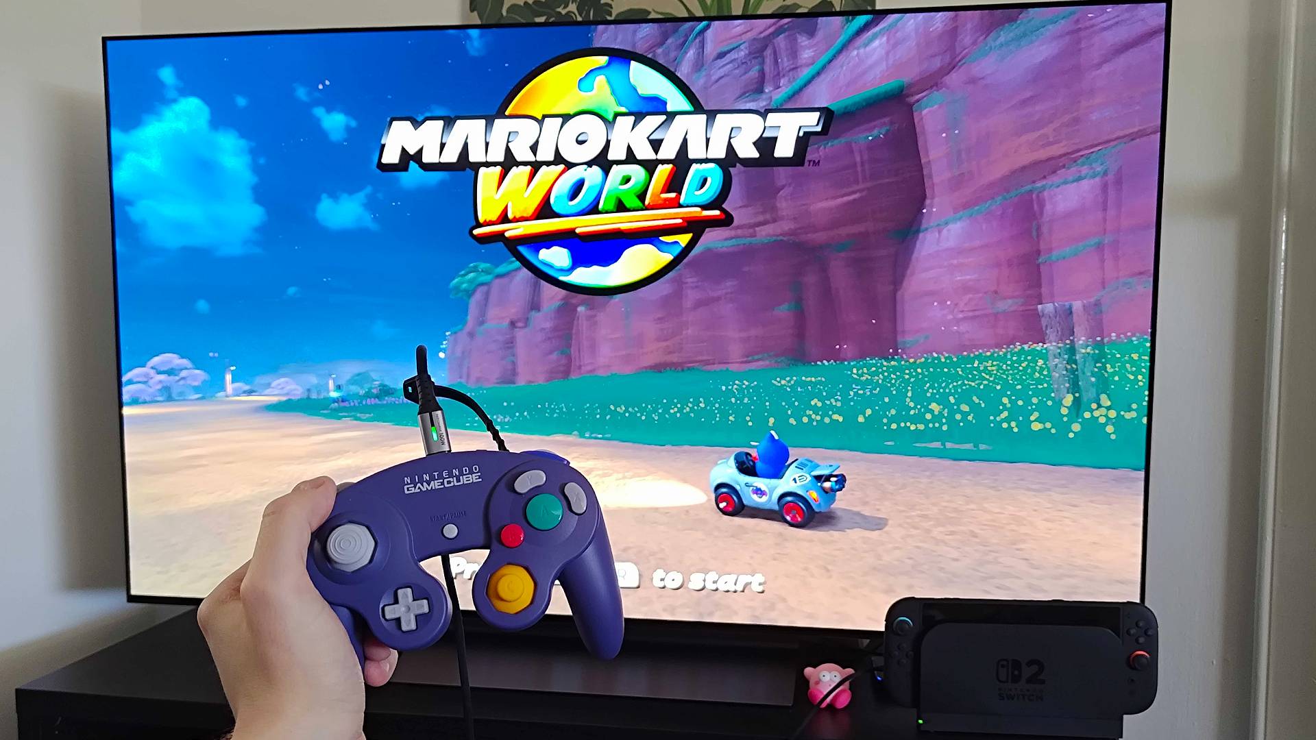 Hand holding Nintendo GameCube controller connected to Switch 2 using USB-C cable with Mario Kart World title screen on LG OLED C4 TV.