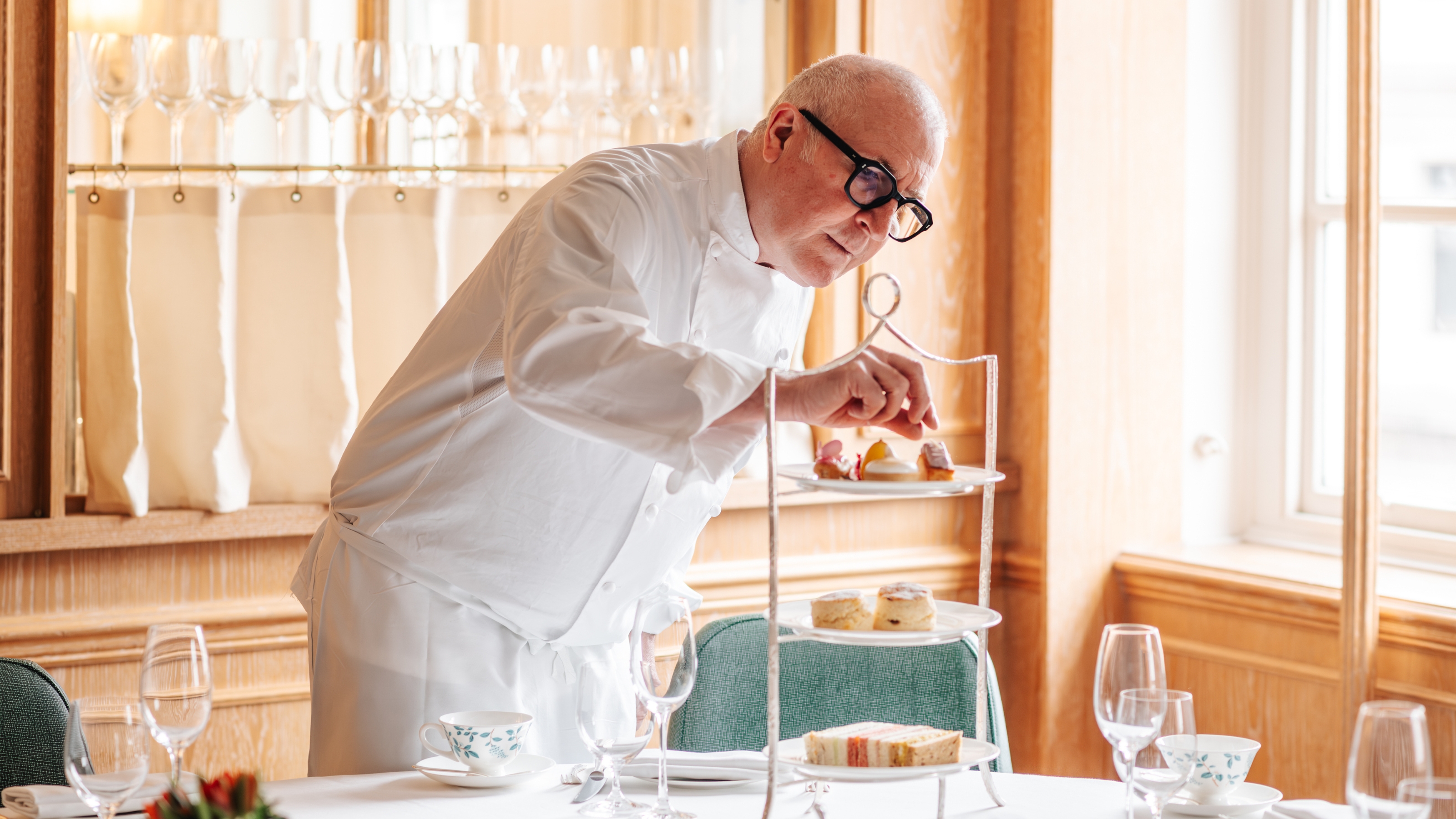 A chef perfects a pastry table arrangement within a plushly decorated tea room with warm wood interiors.