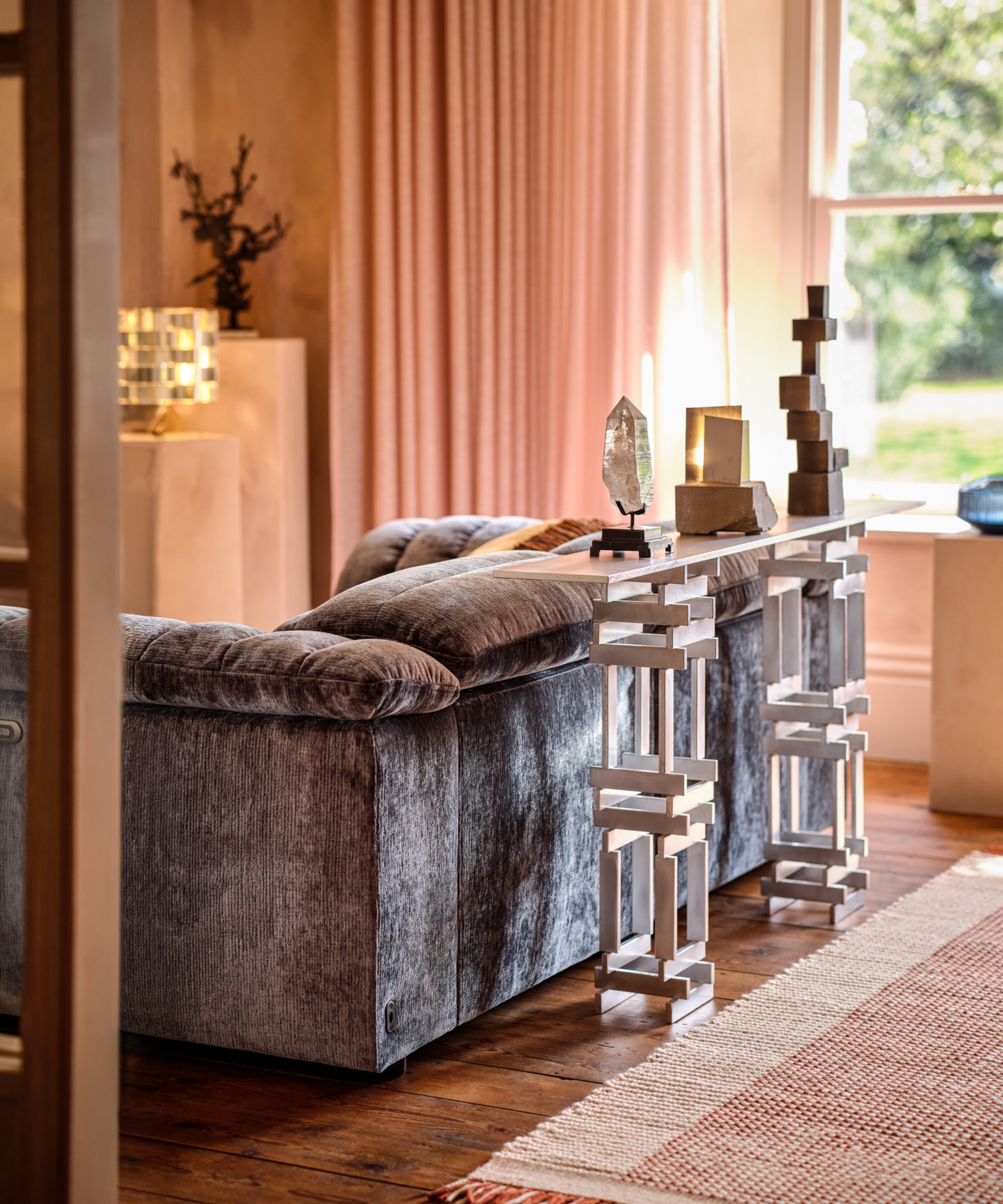 Grey chenille sofa beside a sculptural metal console table with crystal and stone ornaments, pink curtains behind.