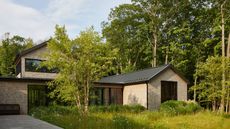 A modern shingle-style house with black-trimmed windows and a metal roof, surrounded by lush green grass and a dense forest.