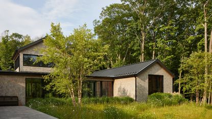 A modern shingle-style house with black-trimmed windows and a metal roof, surrounded by lush green grass and a dense forest.