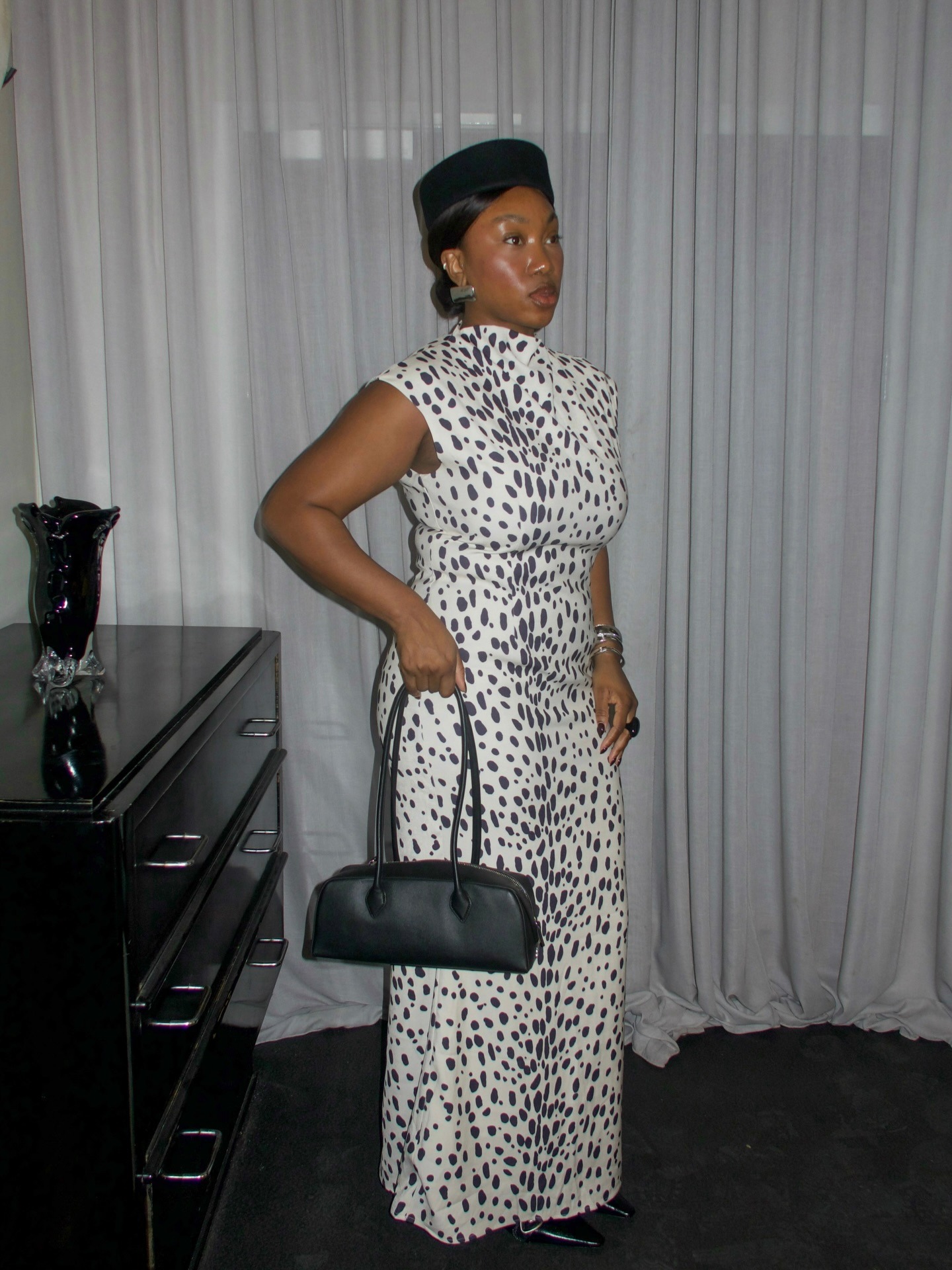 Influencer wearing black pillbox hat, black east-west handbag and long dalmation print dress.