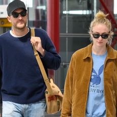 Sophie Turner with Peregrine Pearson at Heathrow Airport wearing a tan jacket, blue Playboy shirt, black leggings, and Ugg mules and carrying a Louis Vuitton bag. 