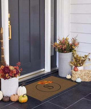 Autumnal front door threshold with grey door and grey tiles, on top of which is a doormat, pumpkins and dried flowers