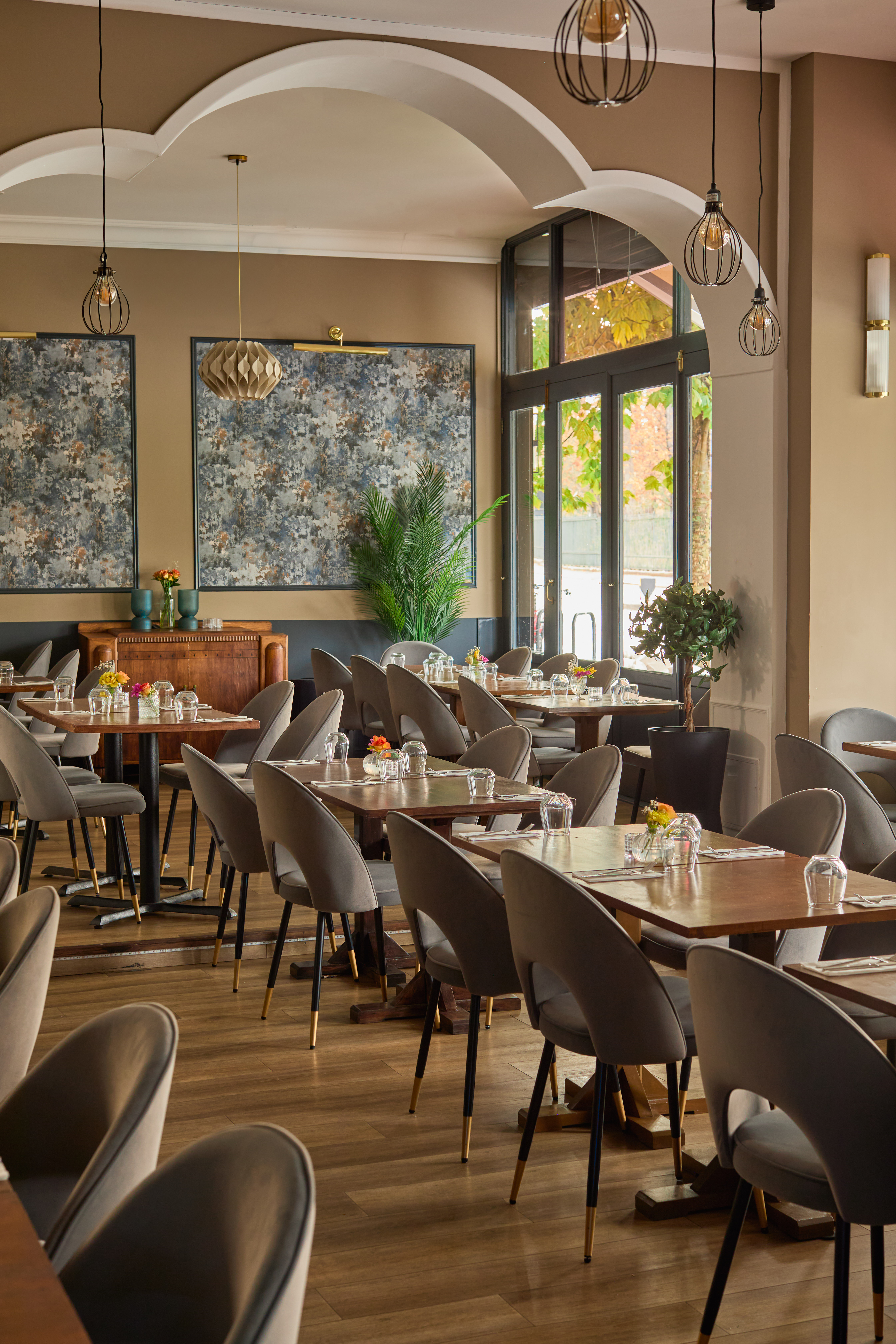 A beige-tinted restaurant decorated with old-glamour style, velvet gray chairs, wooden tables, plants, and abstract paintings.