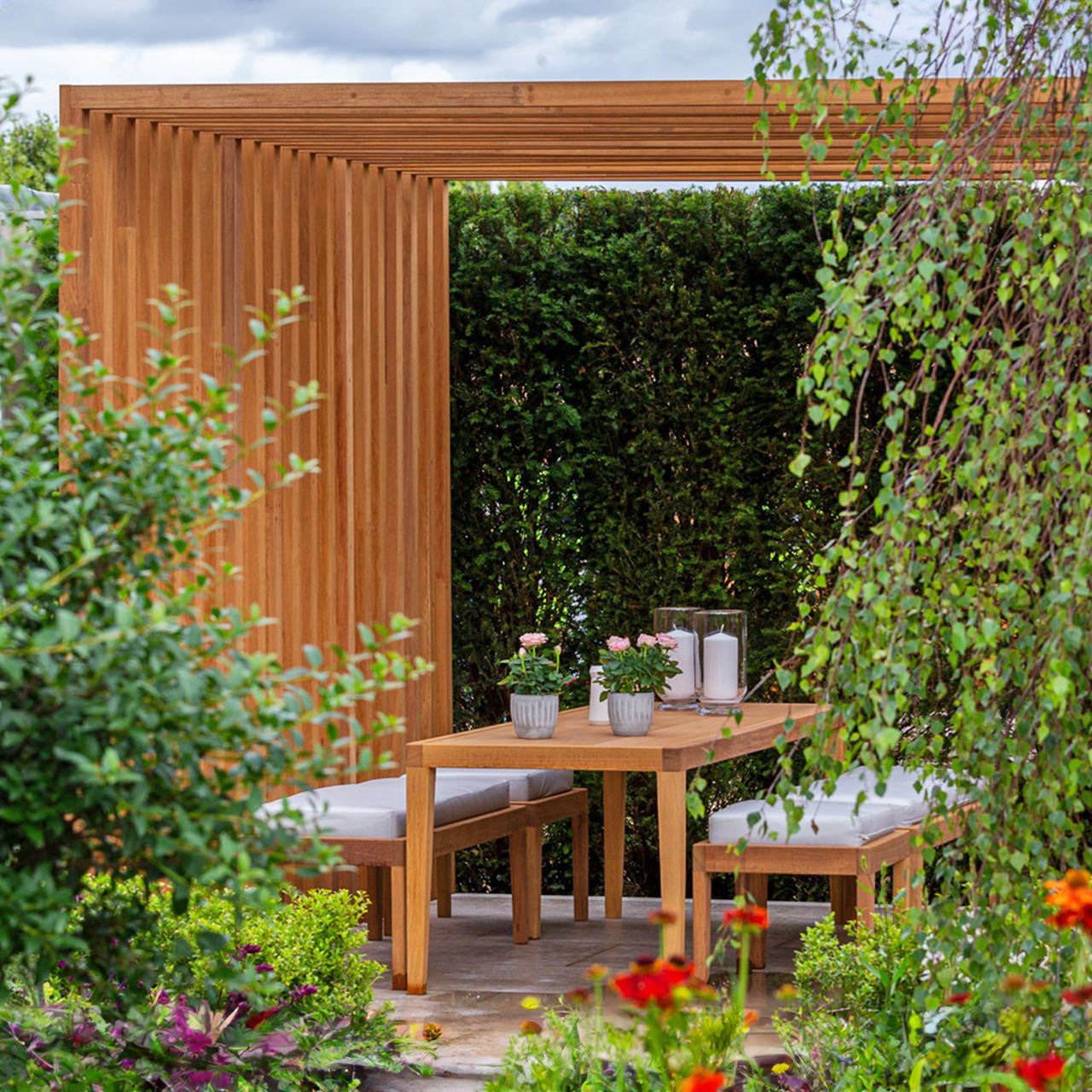 Modern slatted pergola design with dining area and garden furniture, a tree, shrubs and flowers