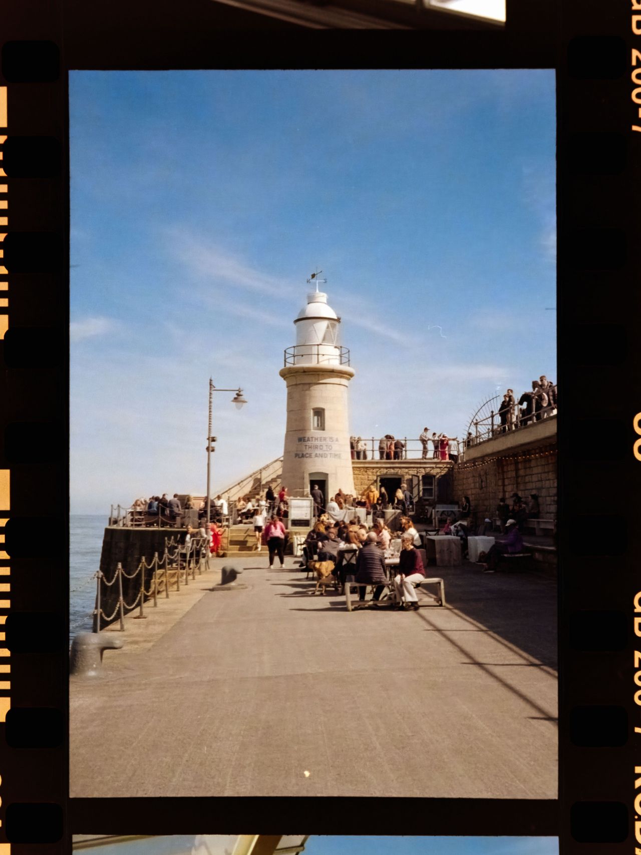 Scanned negative showing a busy seaside pier with a lighthouse, outdoor seating, diners, and people gathered along the waterfront.