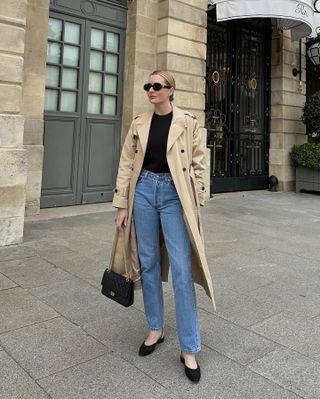 fashion editor Kristen Nichols poses on the streets of Paris wearing an stylish outfit with a trench coat, black sunglasses, black top, classic straight-leg jeans, a Chanel bag, and black low-heel slingbacks