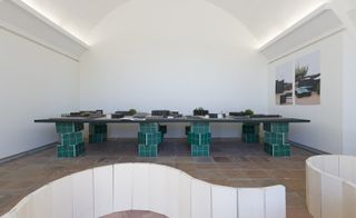 Interior image, red stone floor, white walls, white arched ceiling, strip ceiling lights, long table black top on green brick legs, wall art to the right, white curved walls to the front of the shot