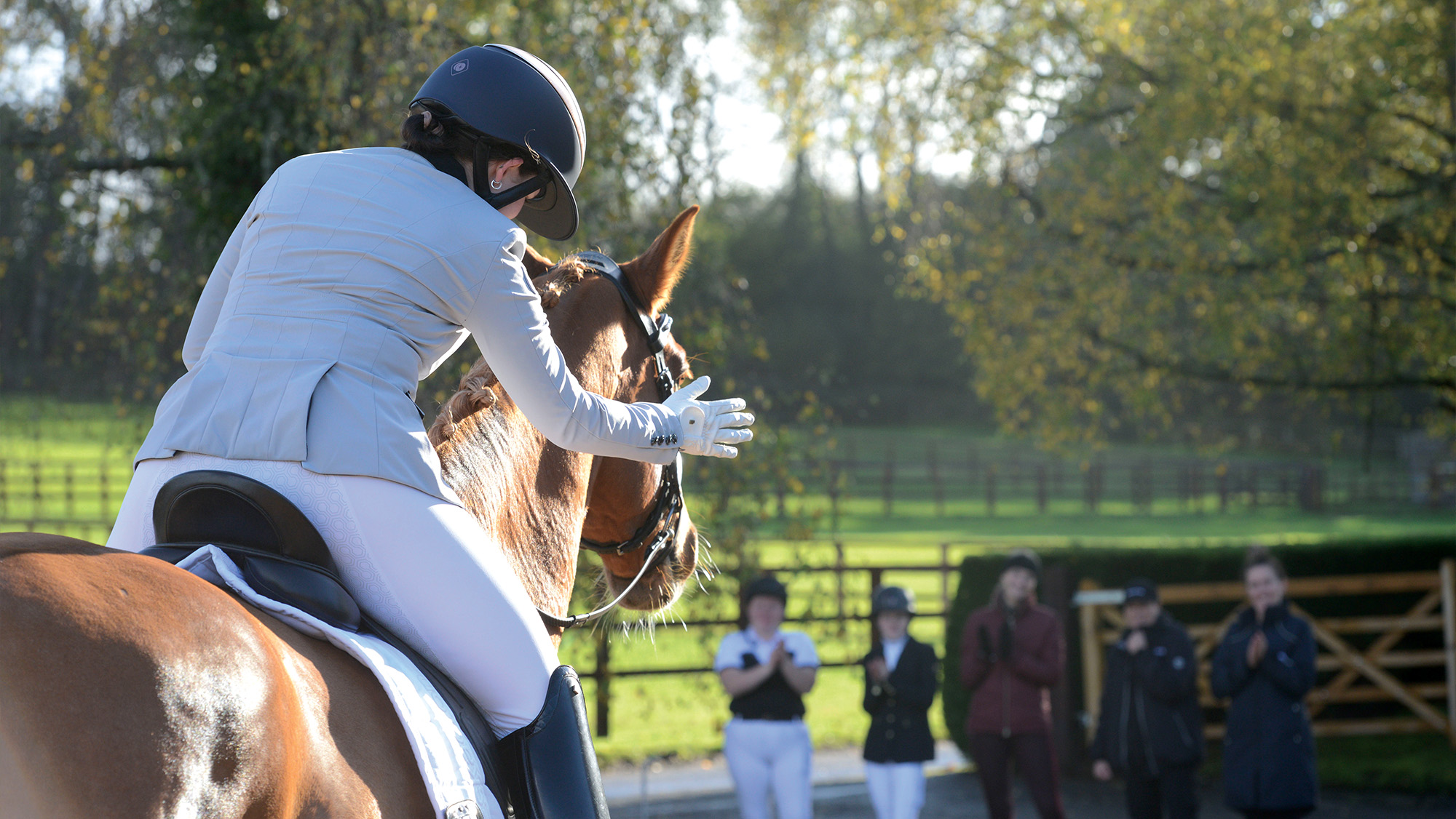 Rider leaning down to pat horse after moving up a level, spectators clapping in the background