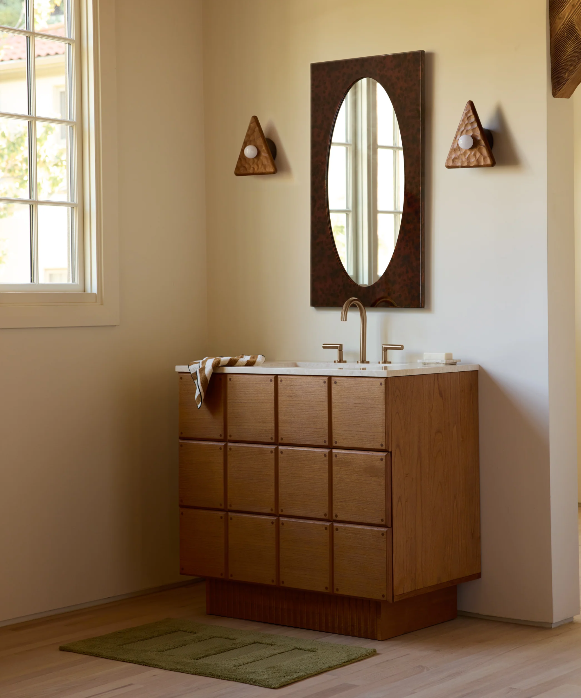 a bathroom vanity with an oval mirror, two triangular wall sconces, and a green bathmat on the floor